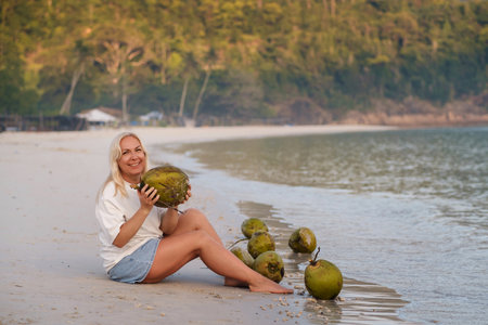 A woman enjoys a tropical beach holding a coconut in her hands, sitting at dawn next to fresh coconuts on a quiet coastline. Redang Island. Malaysiaの写真素材