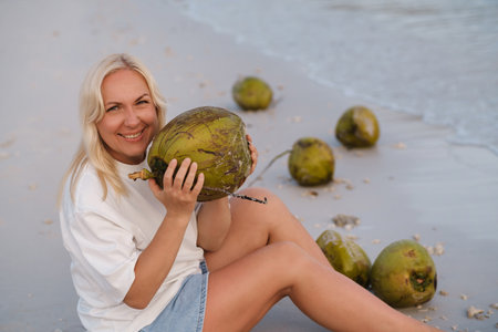 A woman enjoys a tropical beach holding a coconut in her hands, sitting at dawn next to fresh coconuts on a quiet coastline. Redang Island. Malaysiaの写真素材