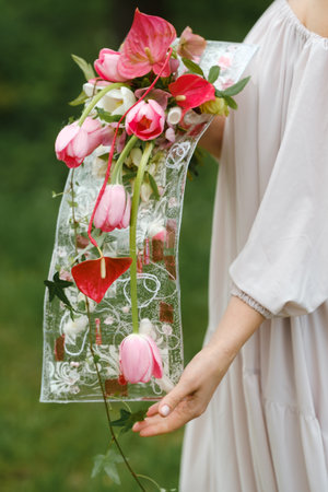 Wedding bouquet with pink tulips and red anthuriums held by a person wearing a light dress in a lush outdoor setting during a sunny dayの写真素材