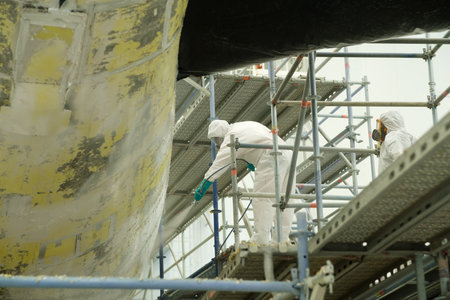 Two workers in white protective suits apply paint to a large ship while using scaffolding at a shipyard, surrounded by cloudy skies and various paintings tools in the background.の写真素材