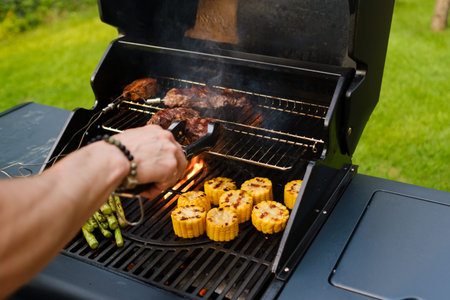 A person is grilling meat and corn on a barbecue in a backyard. The sun is shining, and the grass is...