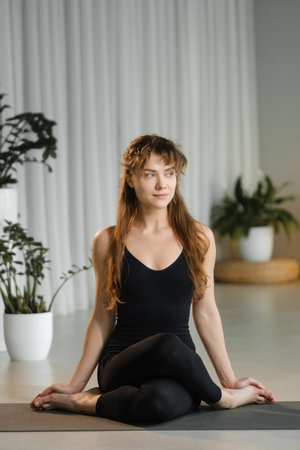 A woman sits on a mat in a yoga pose, concentrating on her breathing. The studio is bright and has...