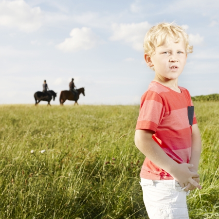 Young blonde boy standing in a grassy field with two horse riders silhouetted on the skyline in the backgroundの写真素材