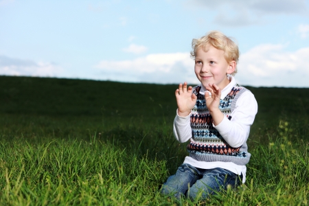 Impish little boy kneeling in a shaft of sunshine in green field smiling as he raises his hands to his chestの写真素材