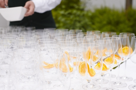 Rows of empty flutes and glasses being prepared with slices of orange before being filled with...