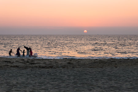 Alleppey beach, India - 14 November 2017: Silhouttes of an Indian family at the beach surprised by a big splashing wave during orange sunset, Alleppey beach, Kerala, Indiaのeditorial素材