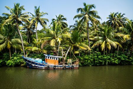 Old ocean fishing boat along the canal Kerala backwaters shore with palm trees at a sunny day between Alappuzha and Kollam, Indiaの写真素材