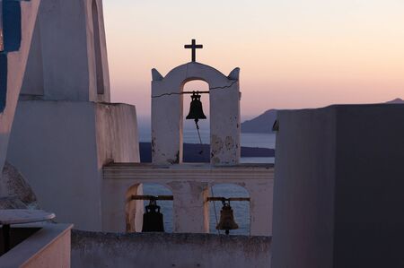Greek bell tower with cross during purple orange sunset along the ocean, Fira, Santorini, Greeceの写真素材