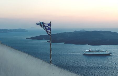 Torn Greek flag along the purple sunset coast with cruise ship in Fira, Santorini, Greeceの写真素材