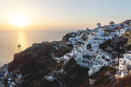 View on Greek village with wind mill on cliffs along the ocean during sunset with sail ship, Oia, Santorini, Greeceの写真素材