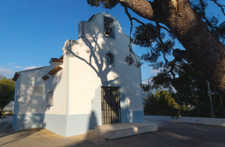 White chapel 'Ermita de Sant Vicent' sunlit with shadows from big old tree in La Nucia, Costa...
