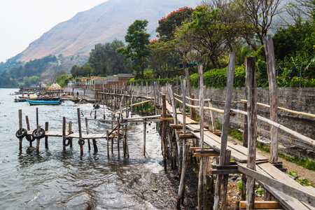 Narrow wooden bridge and docks along lake Atitlan and steep mountain at the coast of Santa Cruz la Laguna, Guatemalaの写真素材