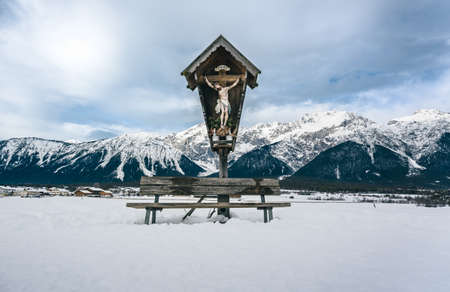 Traditional Austrian Jesus cross with bench in snow winter landscape along the mountains 'Miemingerkette', Mieming Tirol, Austriaの写真素材