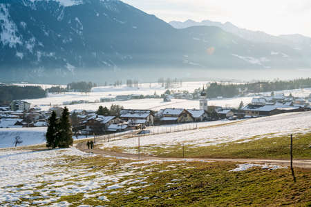 Sunlit village in the mountains with melting snow uncovering the grass meadow, Wildermieming, Tirol, Austriaの写真素材
