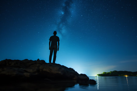 Generative AI : Low angle of silhouette of anonymous tourist standing with light torch on head on cliff against glowing starry sky at nightの素材
