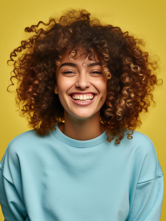 Photo of beautiful young woman with curly hair embraces herself tilts head feels cold during winter evening wears orange jumper isolated over blue background has broad sm : Generative AIの素材