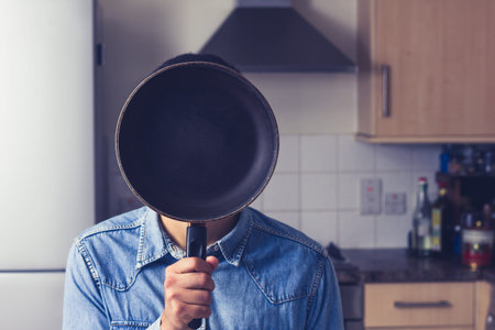 Man is hiding his face behind a frying pan in his neat and tidy kitchen