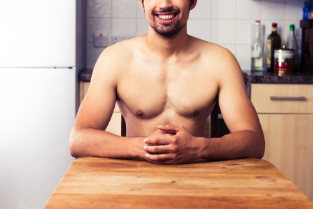 Naked man is sitting at table in kitchen with hands foldedの写真素材