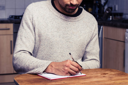 Young man at kitchen table is writing in notebook
