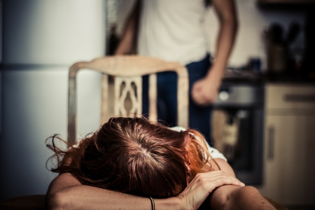 Crying woman at table with abusive husband in background Crying woman at table with abusive husband in background