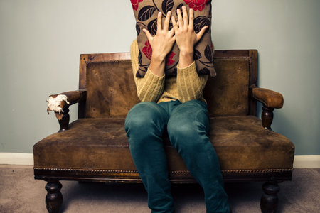 Young scared man on vintage sofa hiding his face behind a cushion