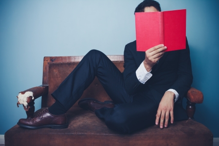 Young businessman is sitting on a sofa and reading a red bookの写真素材
