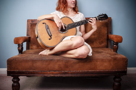Young woman in white dress on sofa is playing guitar