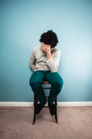 A sad young man is sitting on a high chair by a blue wallの写真素材