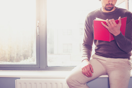 Young man sitting on window sill and reading a red bookの写真素材