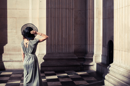 Young woman wearing an elegant dress and hat is standing in an impressive and majestic hall bathed in sunlightの写真素材