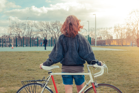 A young woman is standing on the grass in the park with her bicycleの写真素材
