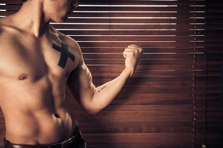 An athletic young man is flexing his muscles by the window in the shade of the venetian blindsの写真素材