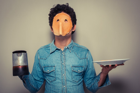 Young man has cut eyeholes in a pancake and is wearing it on his face while holding  a pot of coffeeの写真素材