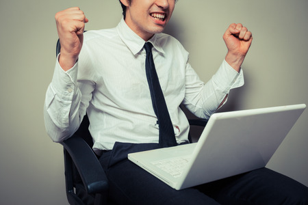 A young businessman is sitting in an office chair and is getting excited about his laptopの写真素材