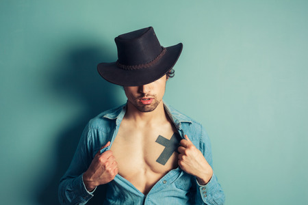 Young cowboy is unbuttoning his shirt to reveal a tattoo on his chestの写真素材