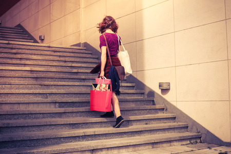 A young woman with shopping bags is walking up the stairs outside at sunsetの写真素材