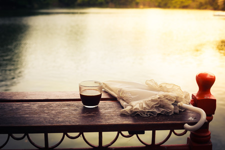 A glass of coffee and an umbrella on a wooden table by a lake in the afternoonの写真素材