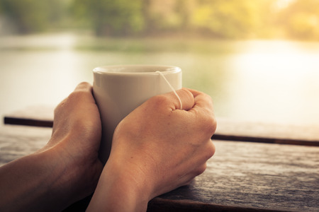 Closeup on a woman's hands holding a cup of tea by a lake in the afternoonの写真素材