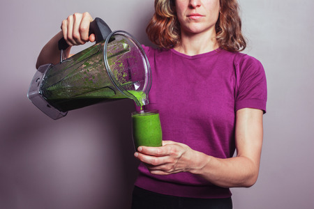 A young woman in a purple top is holding a blender with a green fruit smoothie and pouring it into a glassの写真素材