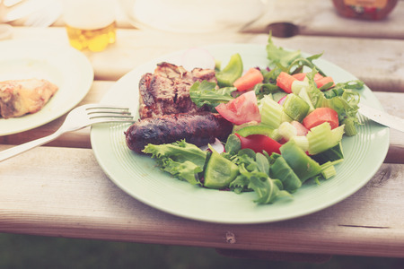 A plate with salad and freshly barbecued meat on a table outside on a sunny dayの写真素材