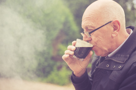 An old man is drinking in a garden with smoke from a barbecue in the backgroundの写真素材