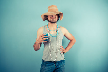 A happy young man wearing a beach hat and pearl necklaces is drinking from a blue cup
