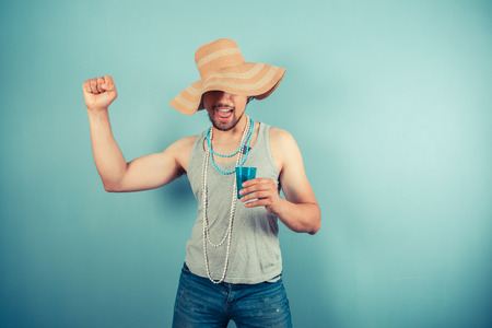 A happy young man wearing a beach hat and pearl necklaces is drinking from a blue cupの写真素材
