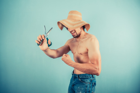 A silly young man wearing a beach hat is playing with scissorsの写真素材