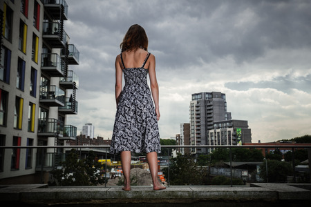 A young woman wearing a dress is looking at a cityscape from a roof on a cloudy dayの写真素材