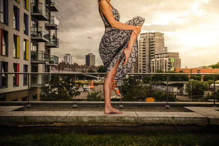 A young woman wearing a dress is raising her leg as she is standing on a roof topの写真素材