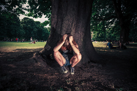 A tired an depressed young woman is sitting under a big tree in the parkの写真素材
