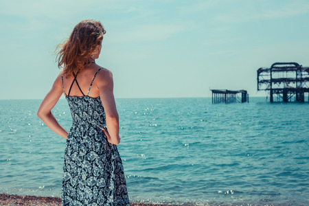 A young woman is standing by the ocean with an old derelict pier in the backgroundの写真素材