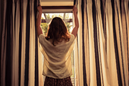 A young woman is opening the curtains at sunriseの写真素材