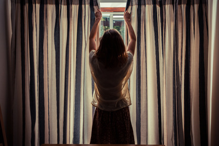 A young woman is opening the curtains at sunriseの写真素材